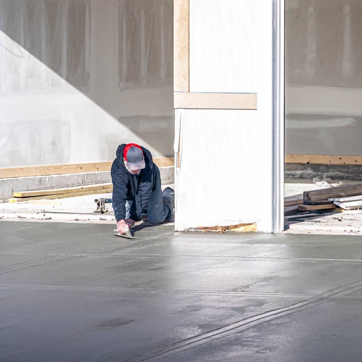 Man kneeling, smoothing wet concrete floor with a trowel in a new building under construction.