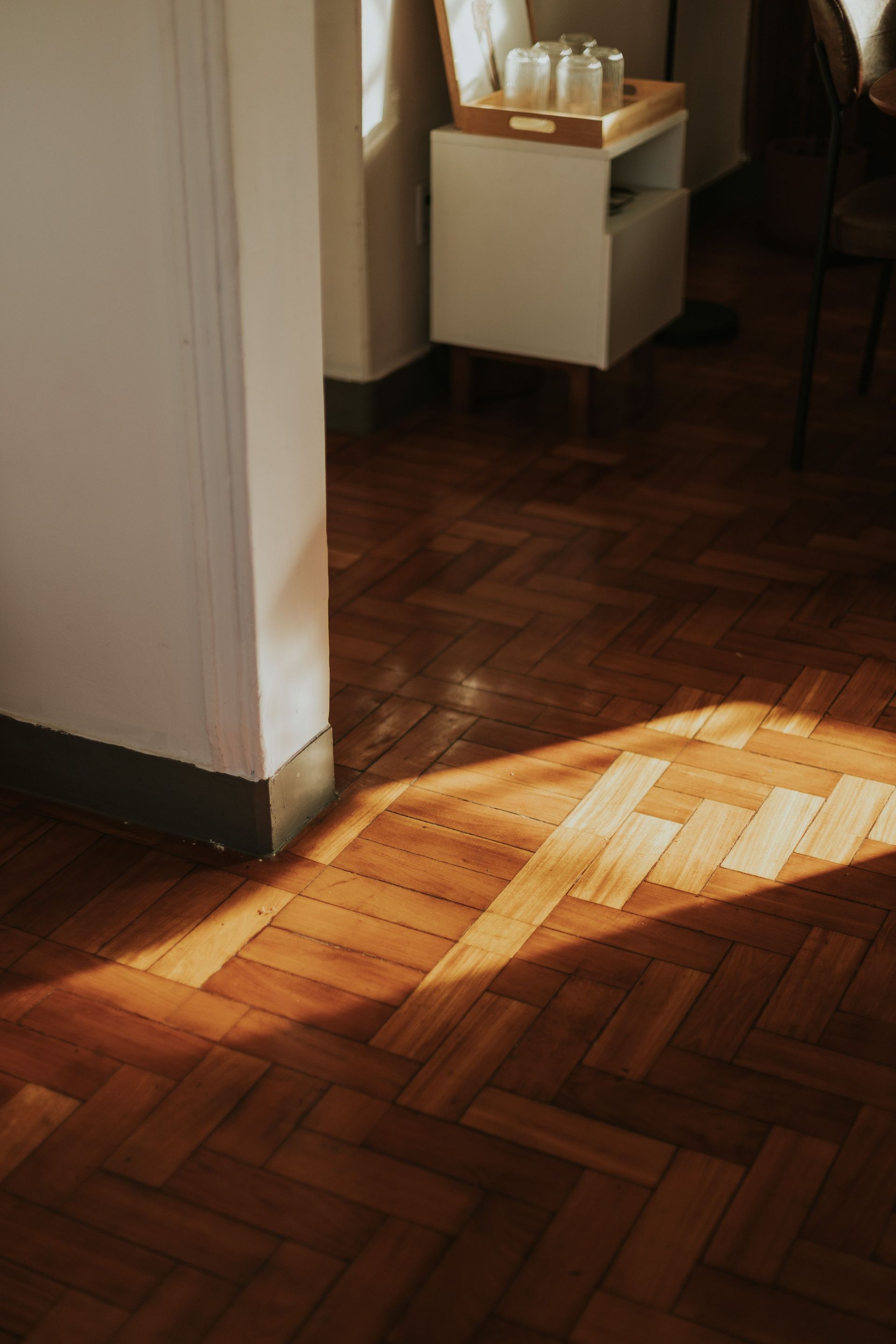 Sunlit wooden floor in a hallway beside a small white cabinet with glasses