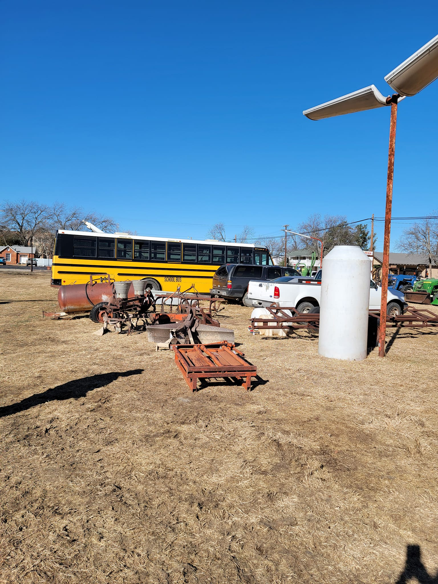 A yellow school bus, farm equipment, and a white storage tank sit in a sunny field under a clear blue sky.