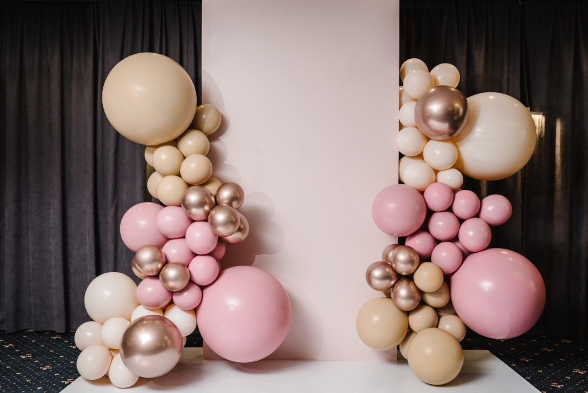 Balloon clusters in blush pink, beige, and gold framing a white backdrop with black curtains