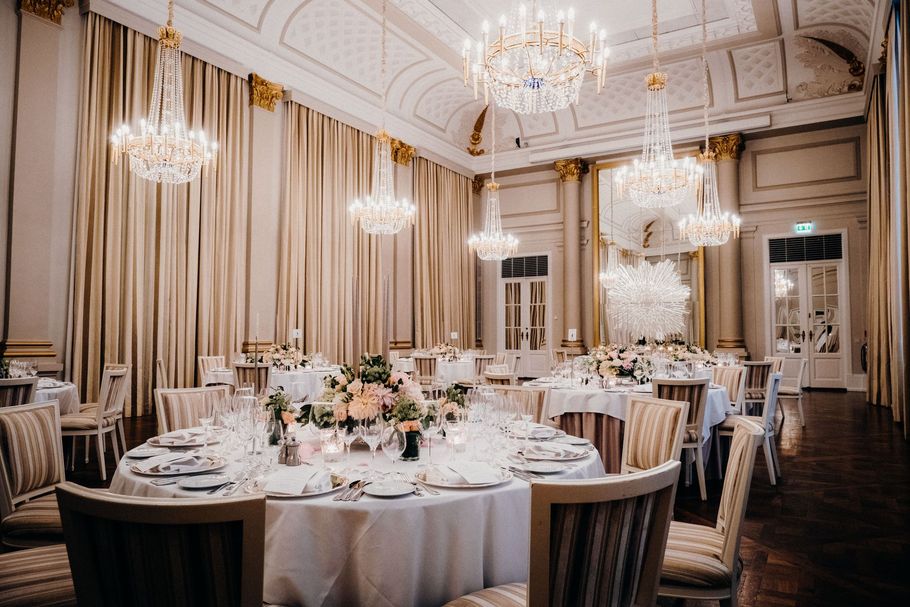 Elegant banquet hall with round, white-clothed tables, chandeliers, and tall draped windows