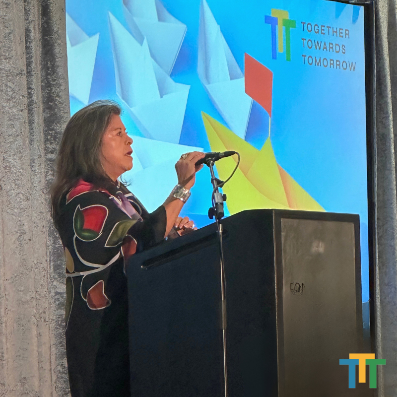 A woman stands at a podium in front of a screen that says together towards tomorrow