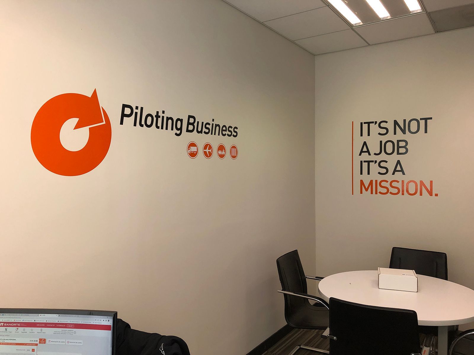 Interior de oficina con vinilos decorativos naranjas: logotipo de la empresa y frase motivadora «No es un trabajo, es una misión». Mesa redonda y sillas.