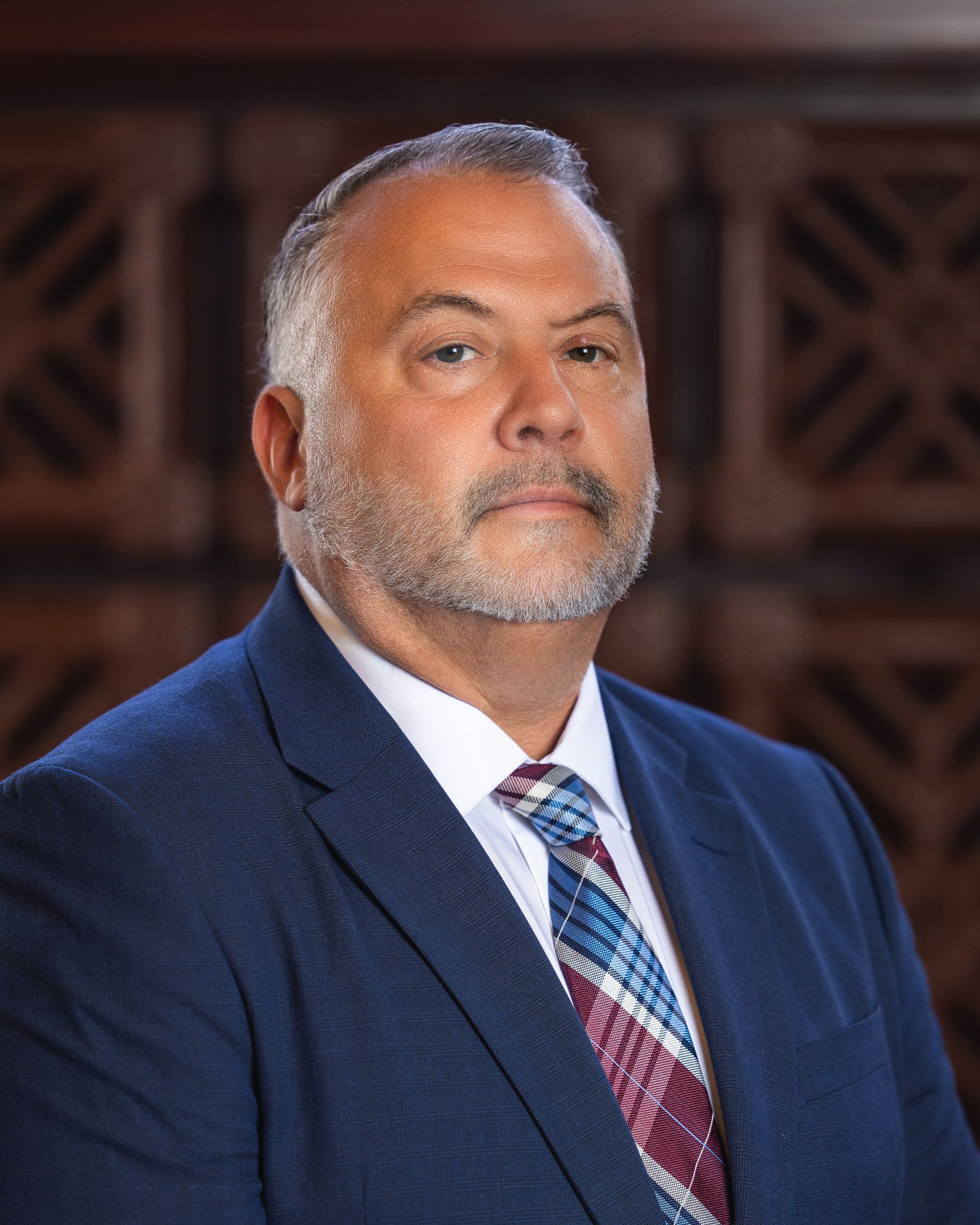 Man in blue suit, gray beard, and patterned tie, looking directly at the viewer. Blurred background.