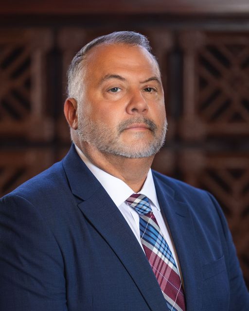 Man in a suit and tie, looking forward, serious expression, against a wood-paneled background.