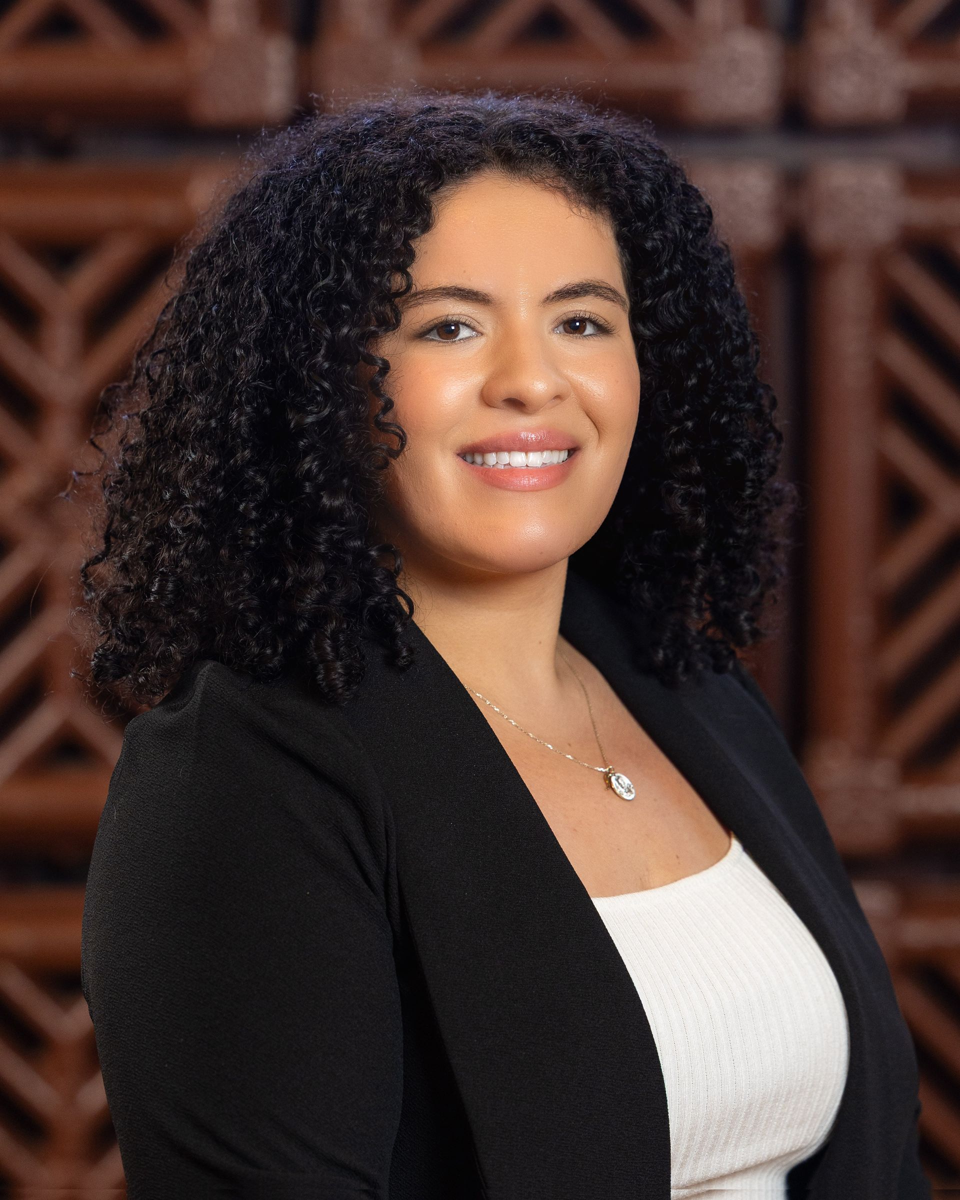 Woman with curly dark hair smiles, wearing a black blazer and white top, set against a wooden background.