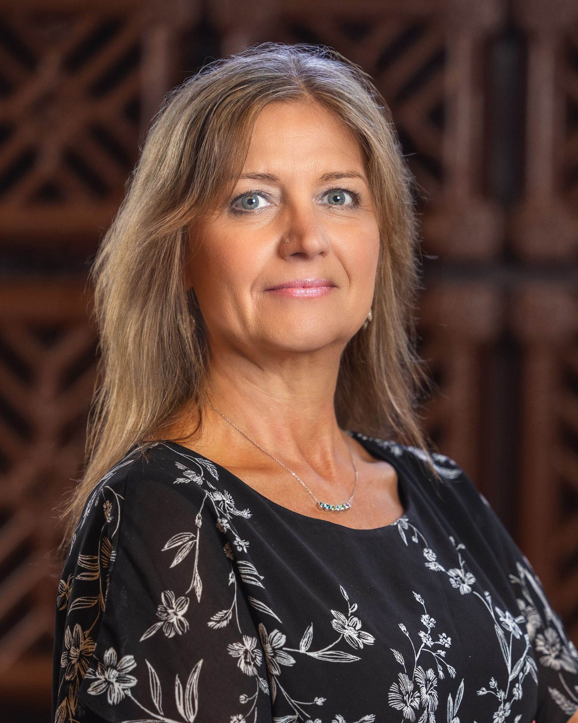 Woman with blonde hair wearing a black floral print top, looking forward, in front of a wooden background.