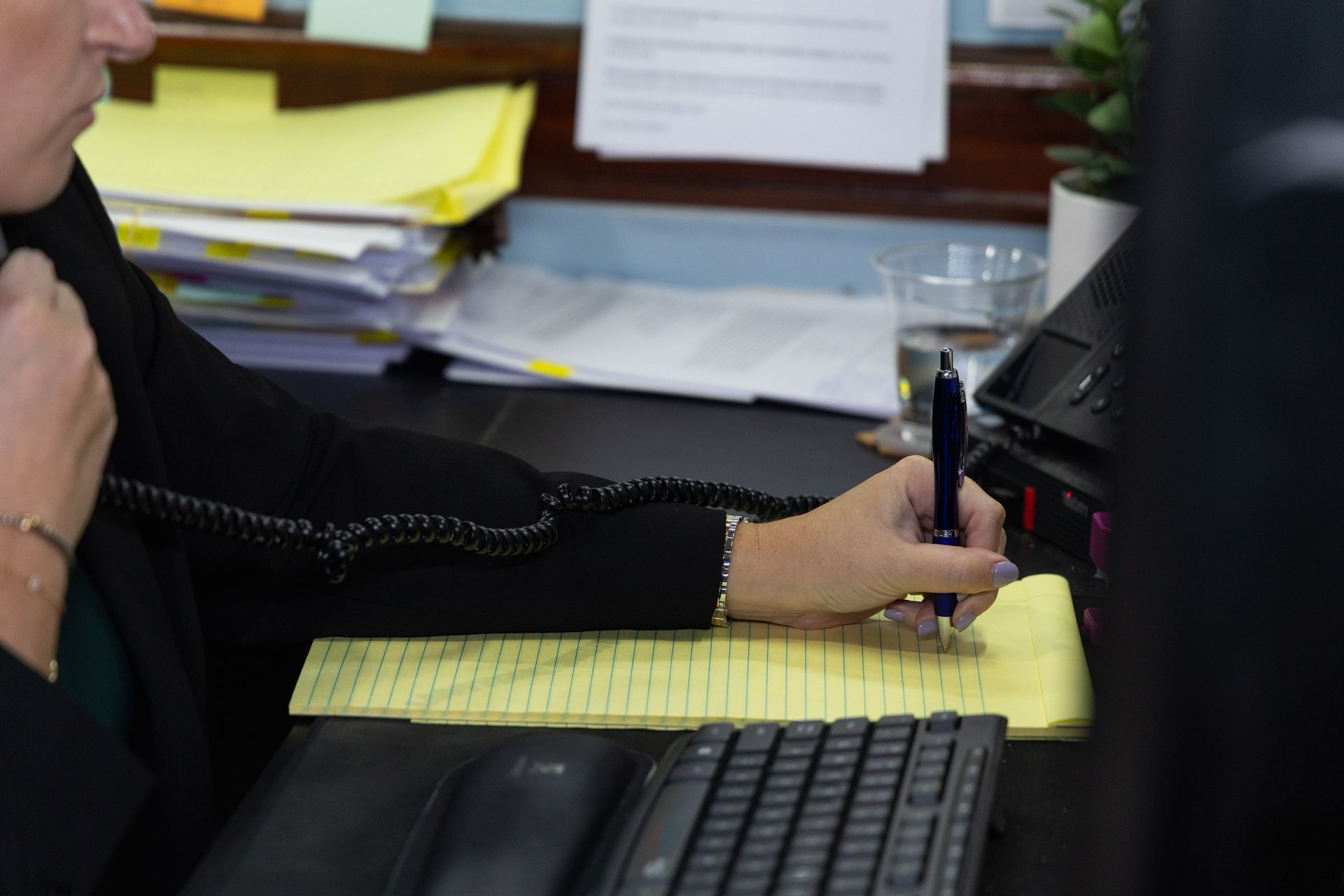 Person writing on a yellow legal pad at a desk with a keyboard and office papers nearby