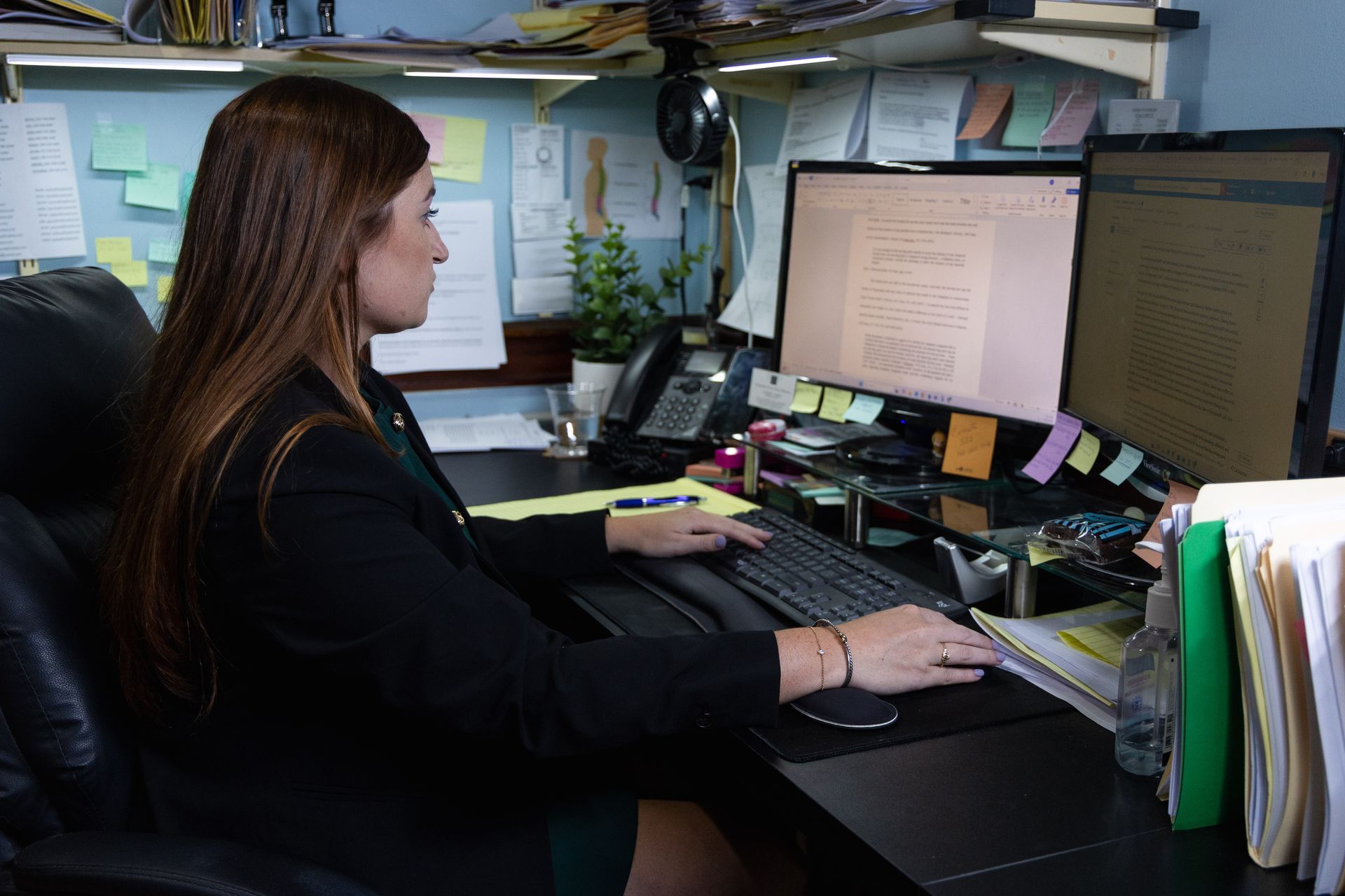 Person typing at a desk with dual monitors in a cluttered office cubicle.