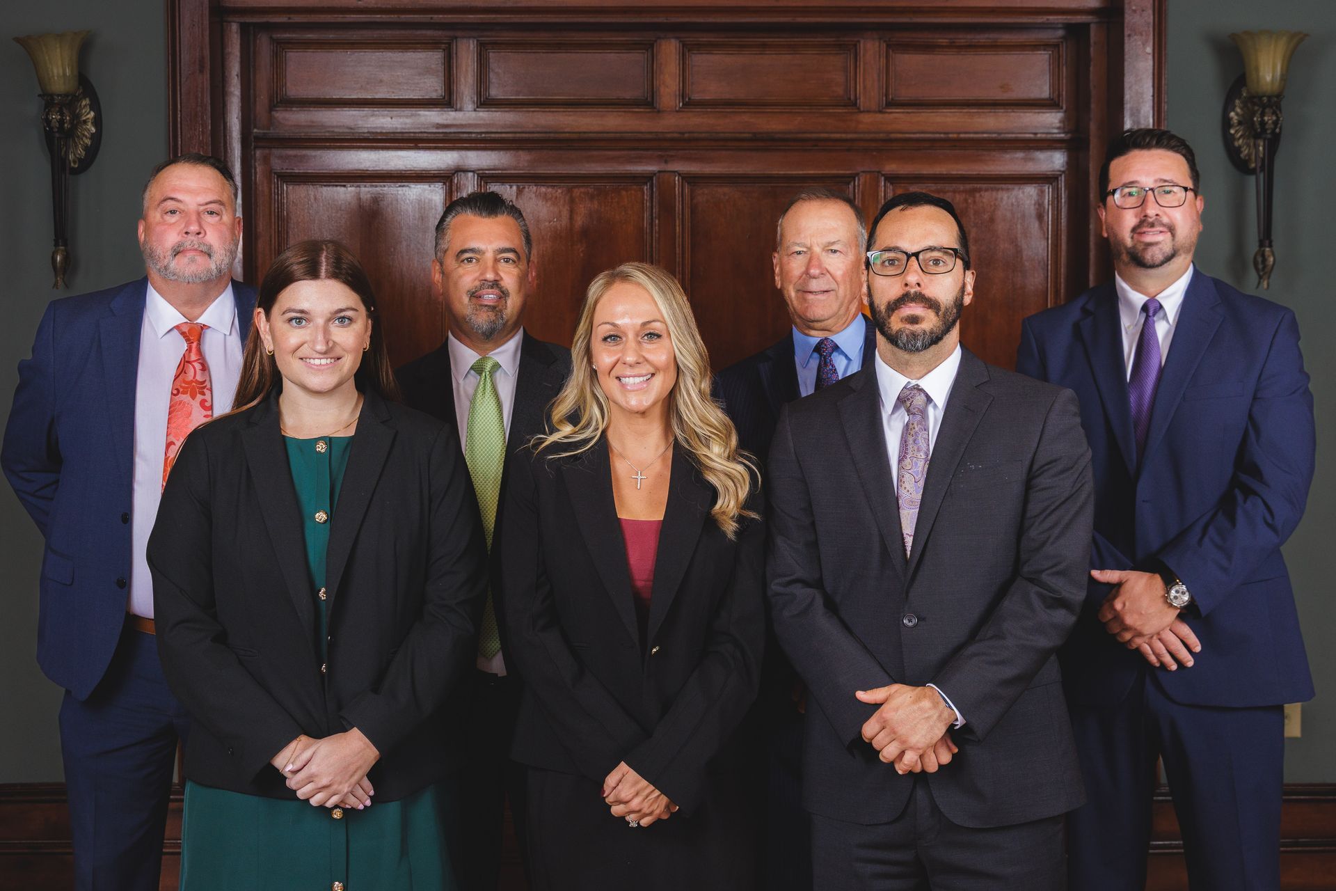 Seven professionals in business attire pose in front of wood-paneled doors.