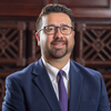 Man wearing glasses, suit, and tie, smiling in front of wood-paneled wall.