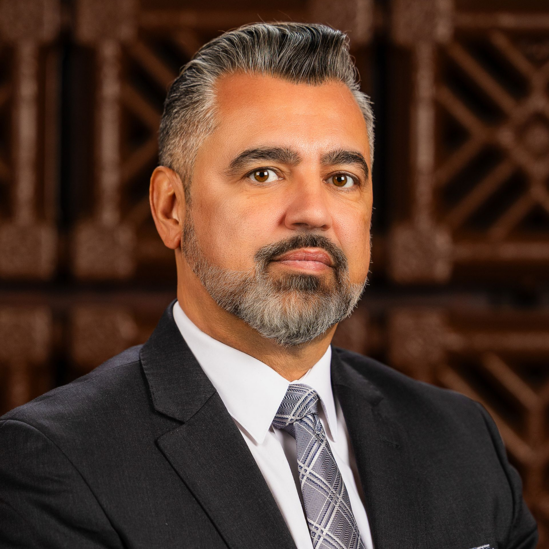Man in suit and tie, against wood background, looking directly at the viewer.
