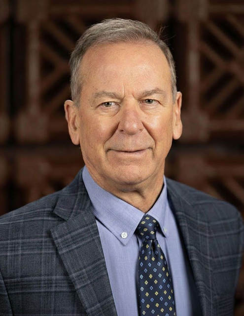 Man in a blue suit and tie, smiling slightly, standing in front of a wooden backdrop.
