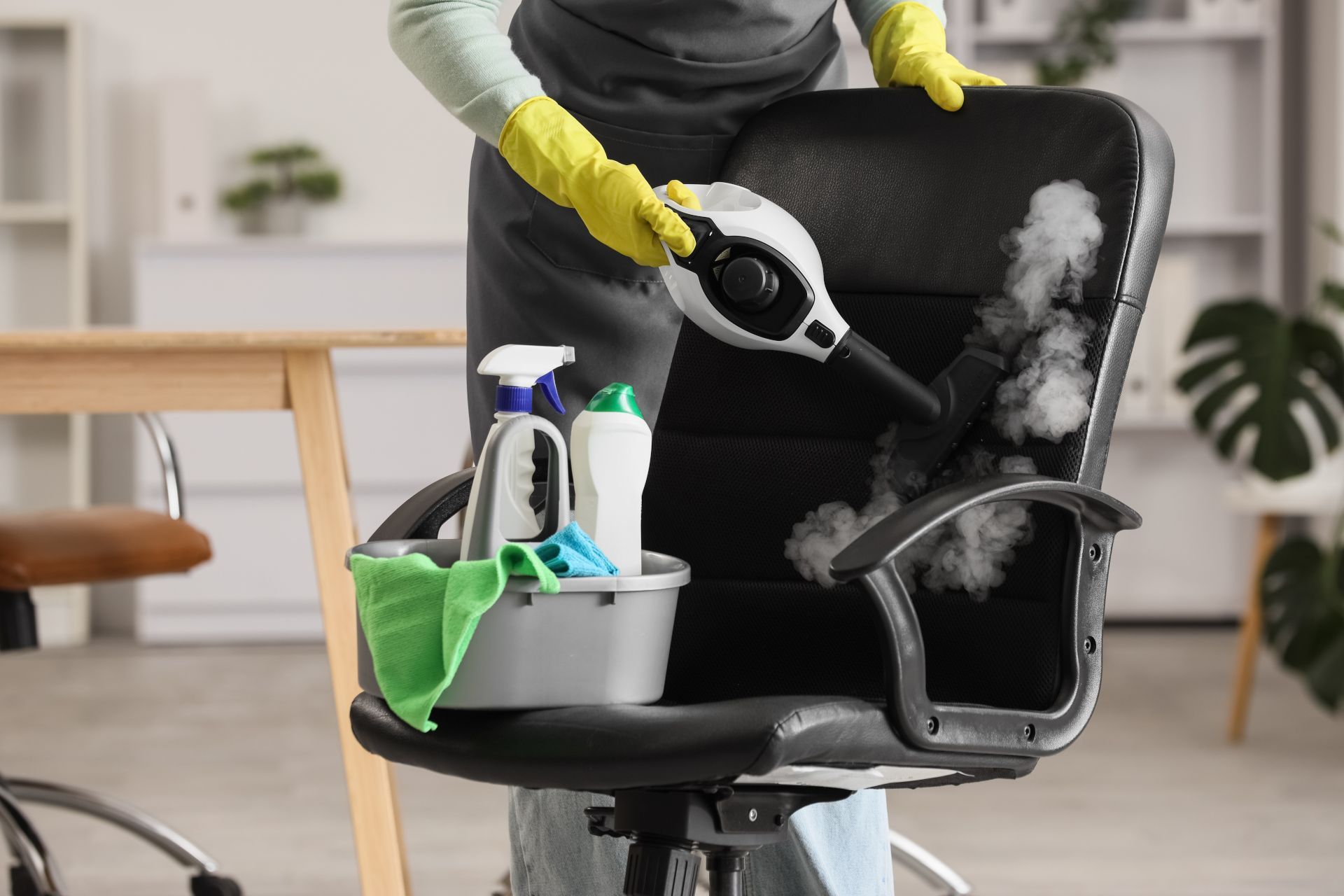 Person cleaning a black office chair with spray cleaner and cloth, holding a steam cleaner in a bright room