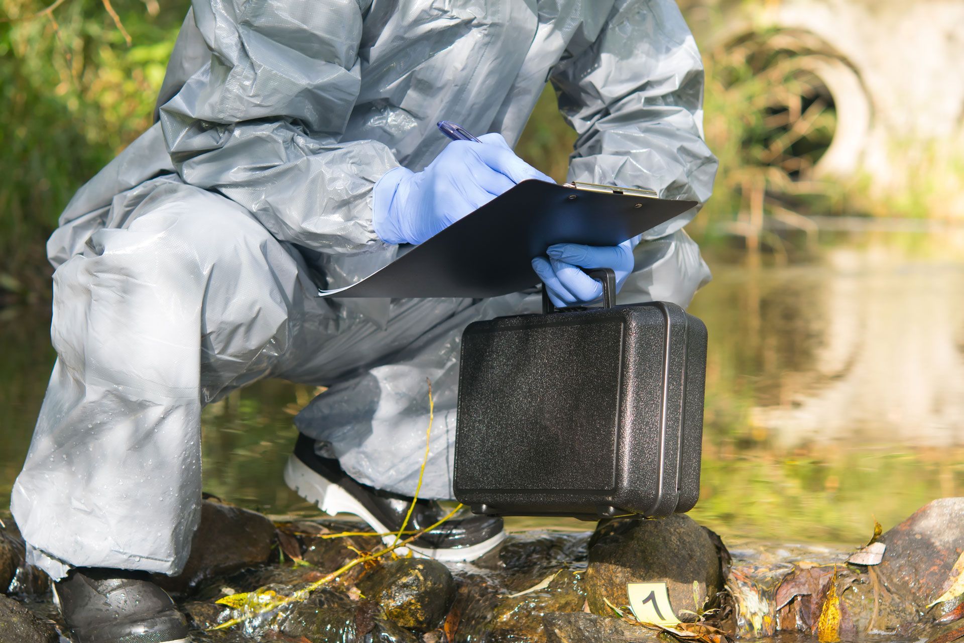 Person in a silver hazmat suit and blue gloves collecting a black sample case by a riverbank