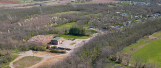 Aerial view of a building surrounded by trees and a grassy area. A small town is visible in the background.