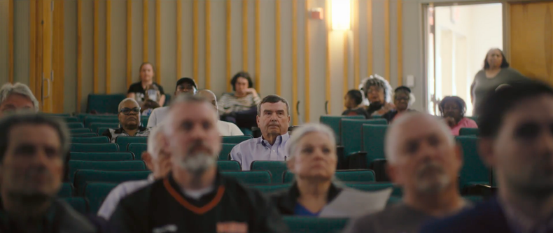 Audience in a meeting room, some looking at the camera. Yellow walls, green seating, and a door open in the background.