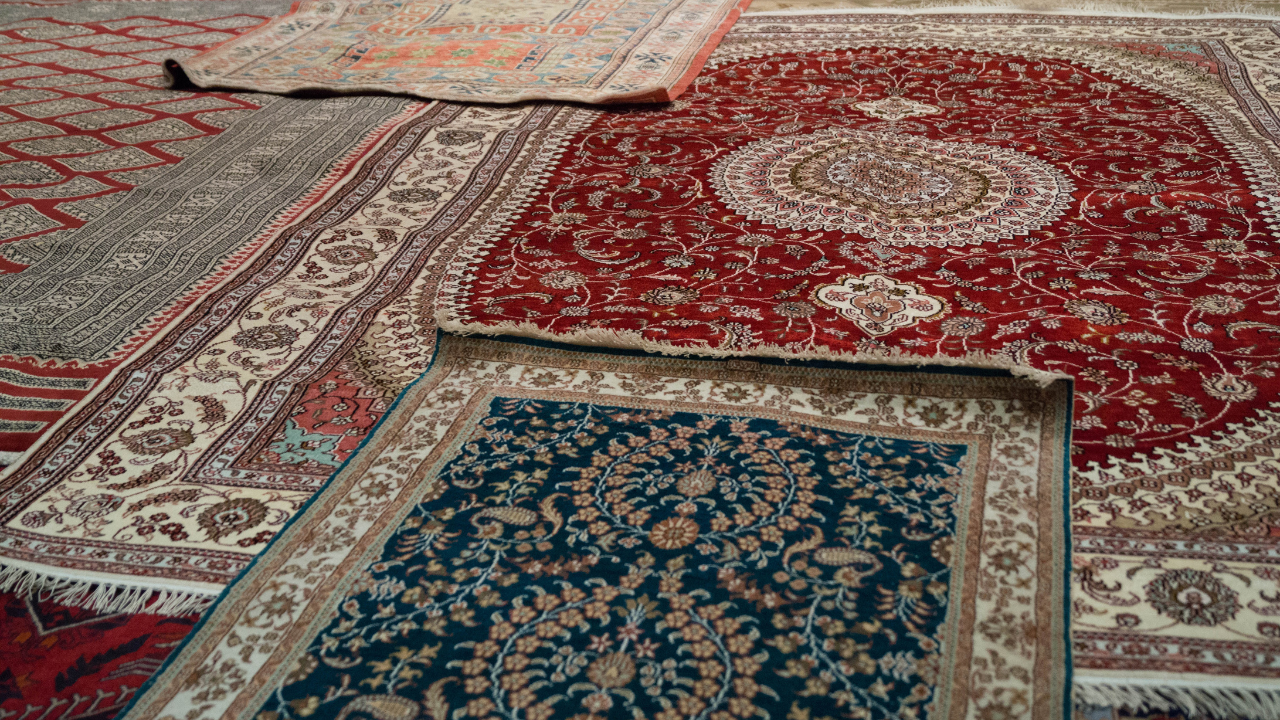 Various ornate rugs, mostly red, cream, and teal, displayed at an angle, showcasing detailed patterns.