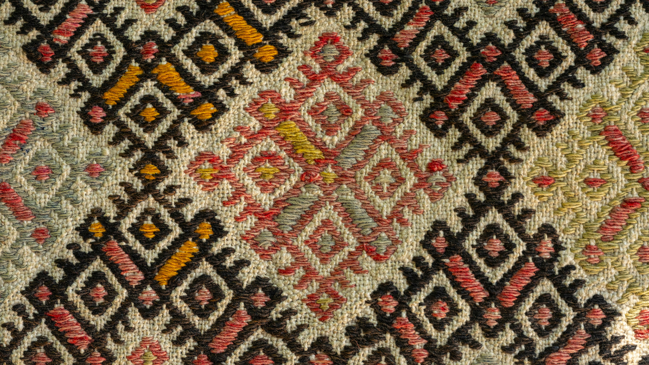 Close-up of a textile with a repeating diamond pattern in black, red, yellow, and white embroidery.