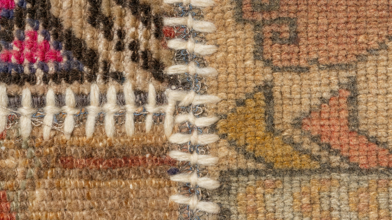 Close-up of a rug, stitched together with white thread; shows various colors, including red, brown, and tan.