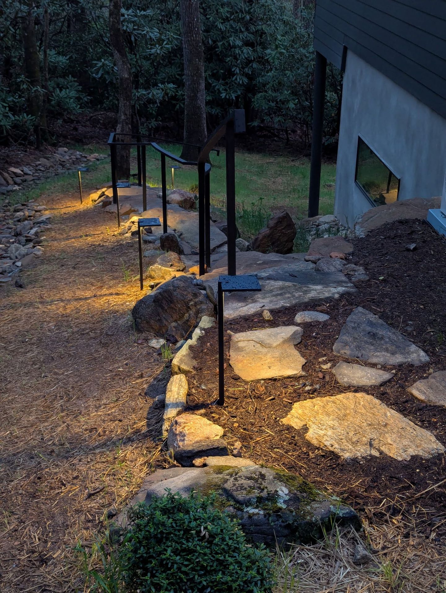 Stone path with warm garden lights beside a building, bordered by rocks and trees at dusk