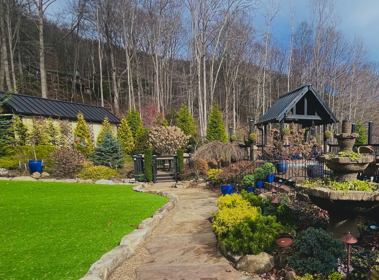 Sunny garden path with green lawn, colorful shrubs, a pergola, and cabins among bare trees
