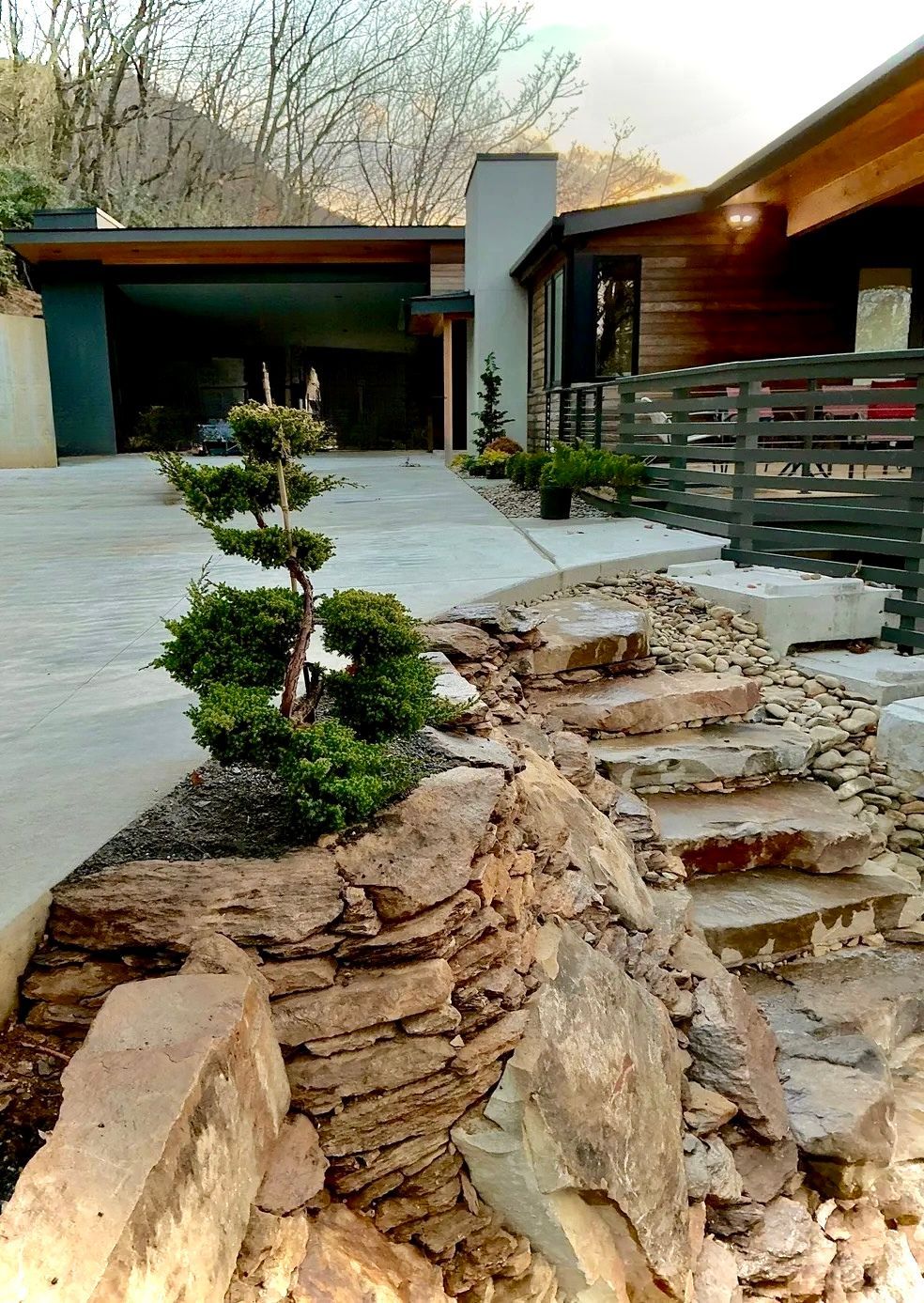 Modern courtyard with stone steps, bonsai, and a concrete driveway beside a house