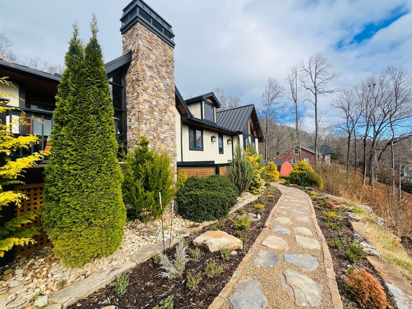 Stone path leading to a house with a tall chimney, evergreen shrubs, and bare trees under a blue sky.