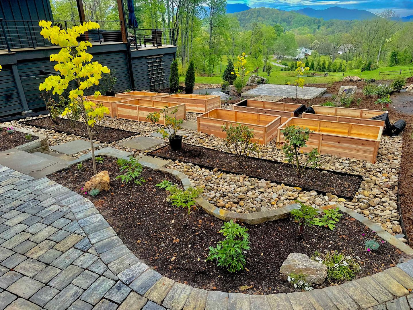 Landscaped garden with stone patio, raised wooden beds, shrubs, and a yellow flowering tree overlooking hills