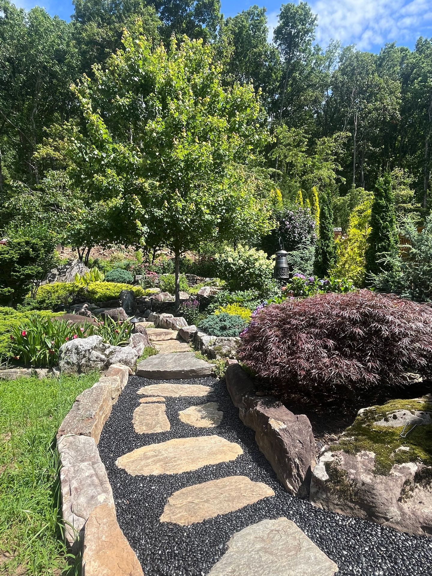 Stone path through a lush garden with green trees, flowers, and a burgundy shrub under bright sunlight