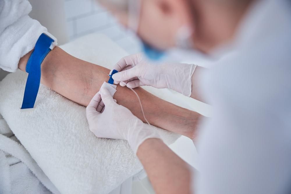 Person in gloves inserting a needle into a patient's arm for IV. White coat, blue tourniquet, close-up.