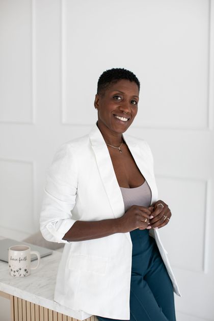 Person in a white blazer and blue pants smiling beside a table in a bright room