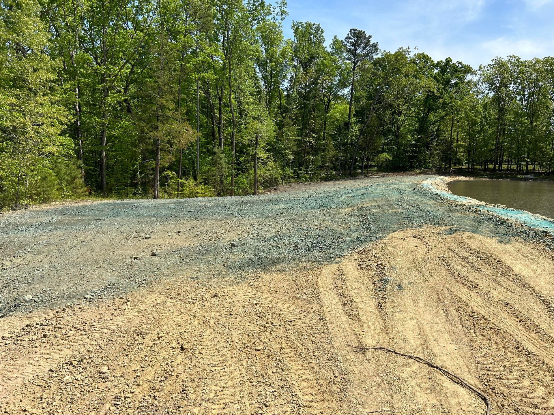 An unfinished construction site with gravel, exposed soil, and tire tracks leading toward a pond near a forest.