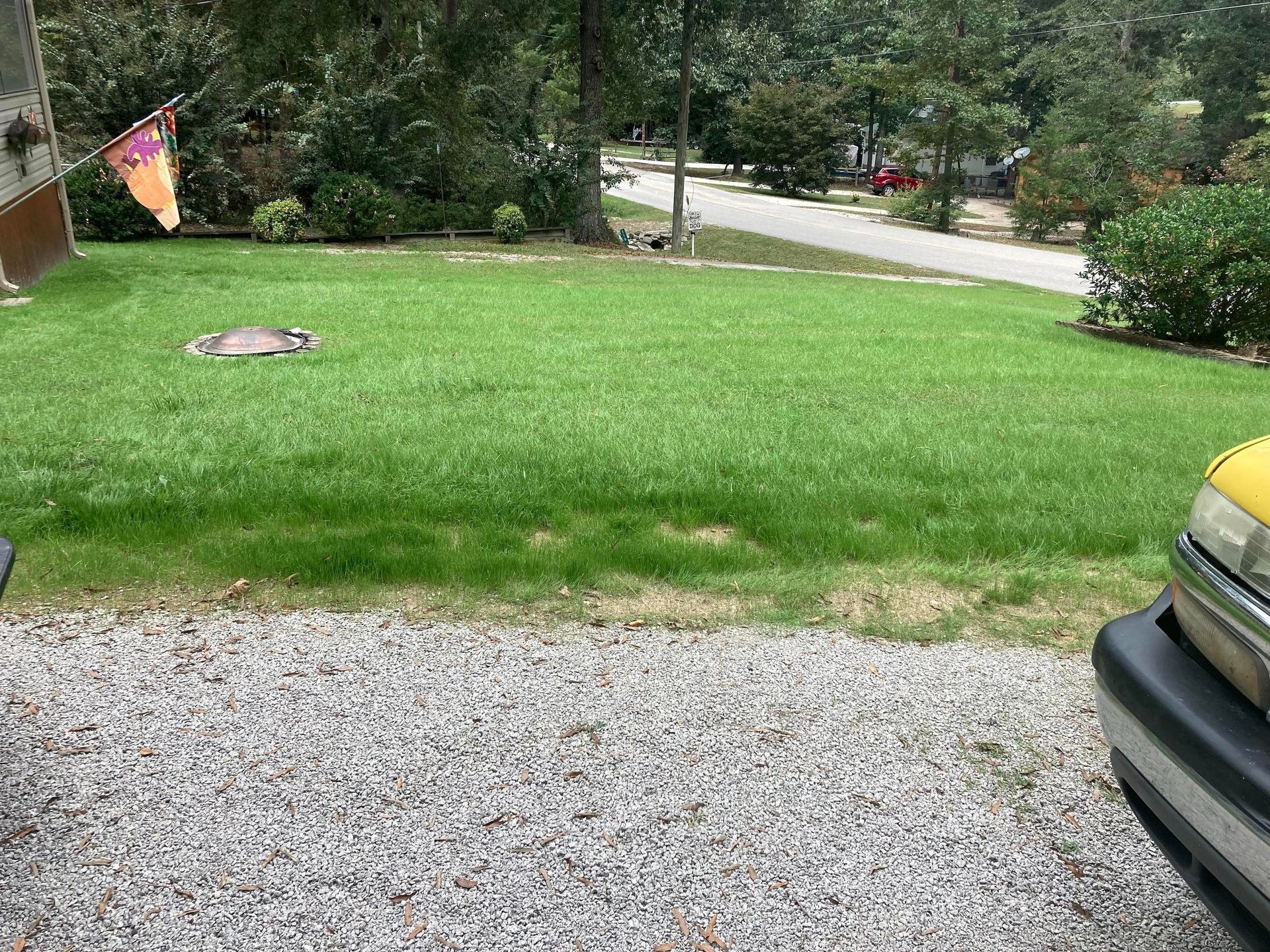 A gravel driveway in the foreground leads to a lush green lawn, with a decorative flag and trees in the background.