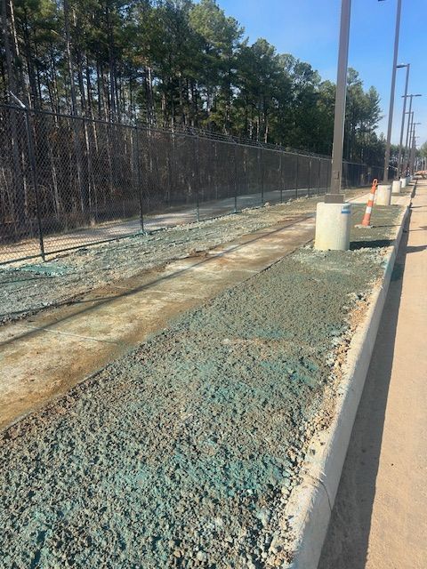 Construction site along a fenced wooded area with a gravel-filled curb and streetlights.