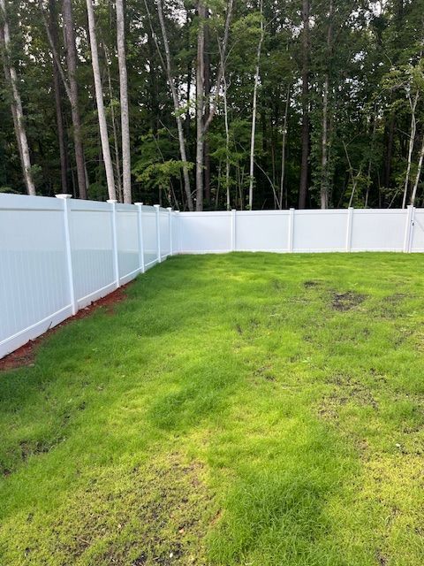 A white vinyl privacy fence borders a grassy backyard, with a line of tall trees in the background.