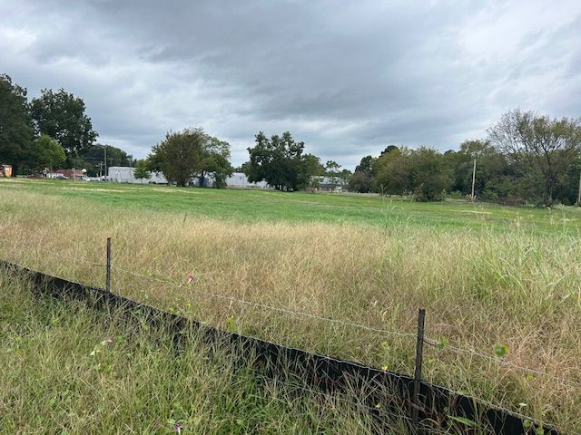 A grassy, open field under a cloudy, overcast sky, bordered by a wire fence and several trees in the distance.