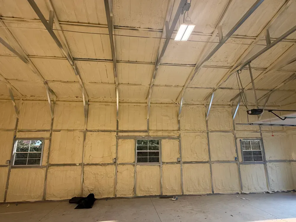 Interior of a building insulated with beige spray foam; metal beams and three windows are visible.