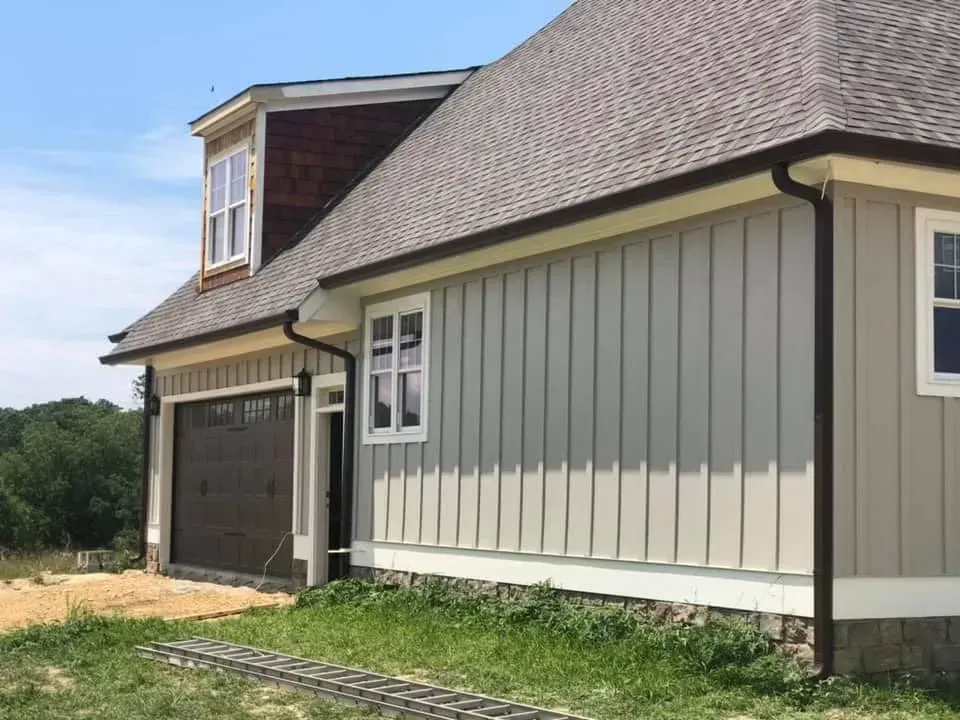 Beige siding on a house with brown trim and roof, a garage door, and white-framed windows.