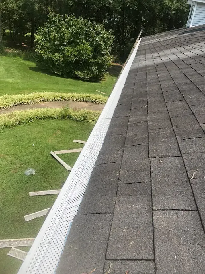 Gutter guard along a dark shingle roof with a green lawn and trees in the background.