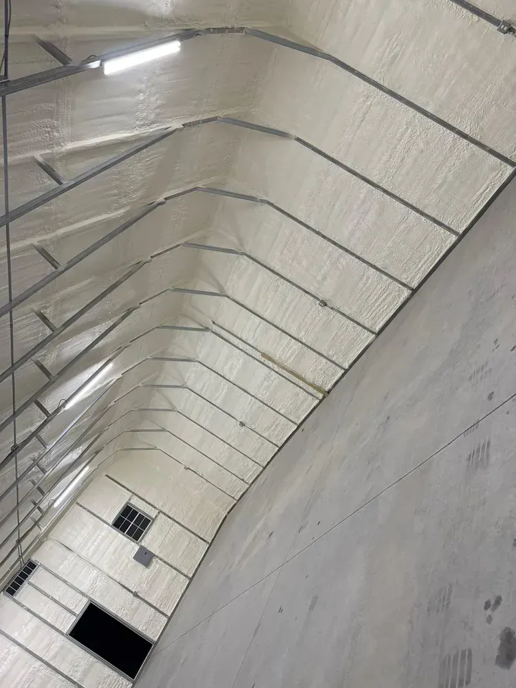 Interior view of a building with spray foam insulation on a curved ceiling with metal beams and a concrete wall.