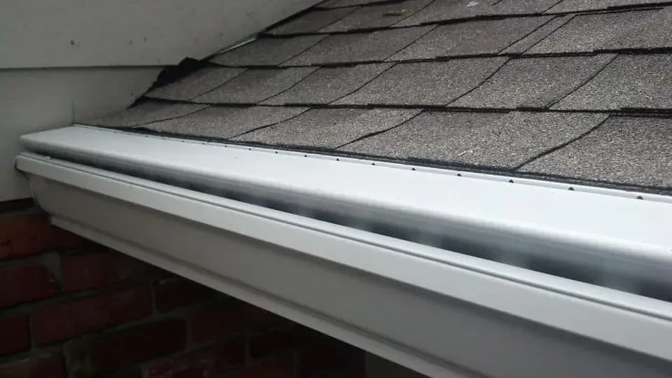 White gutter along a roof edge with gray shingles and a red brick wall.
