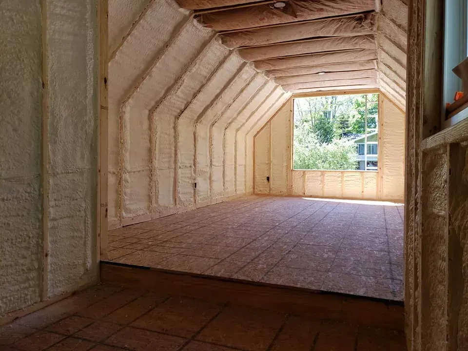 Interior of a room with spray foam insulation on walls and ceiling, a window, and wooden flooring.