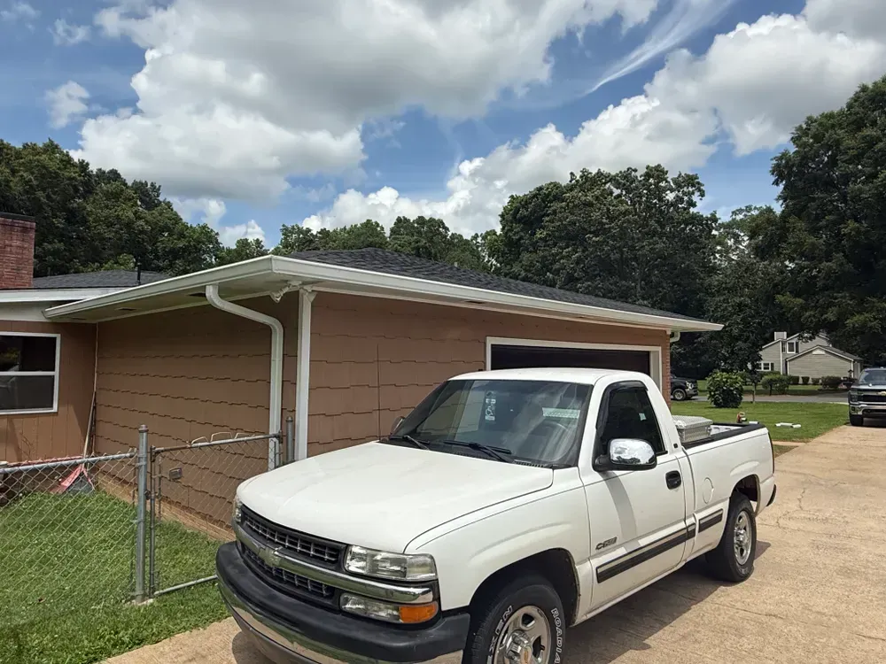 White pickup truck parked in front of a tan building with a dark roof on a sunny day.
