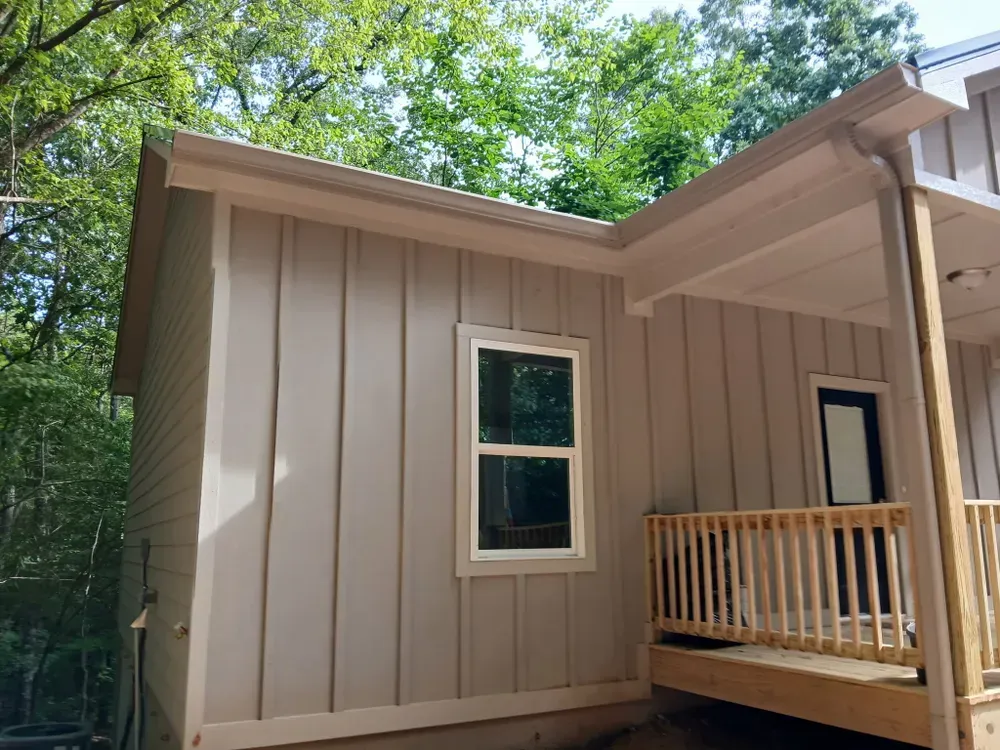 Beige siding on a small cabin with a white-framed window, porch, and a lush green background.