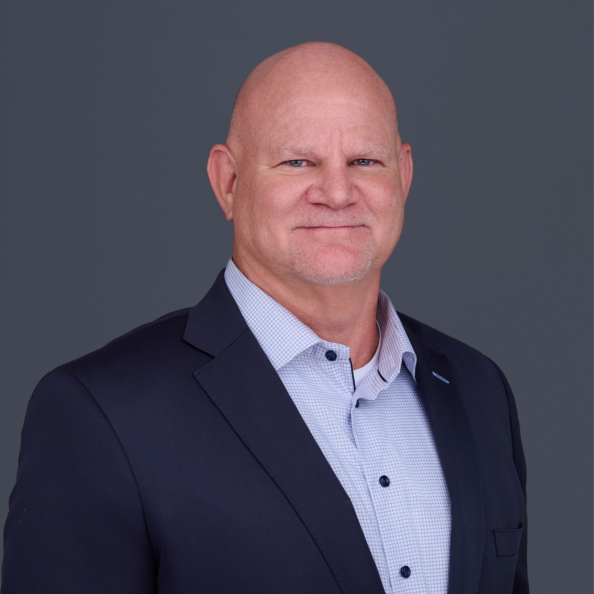 A bald man in a suit and striped shirt is standing in front of a gray background.