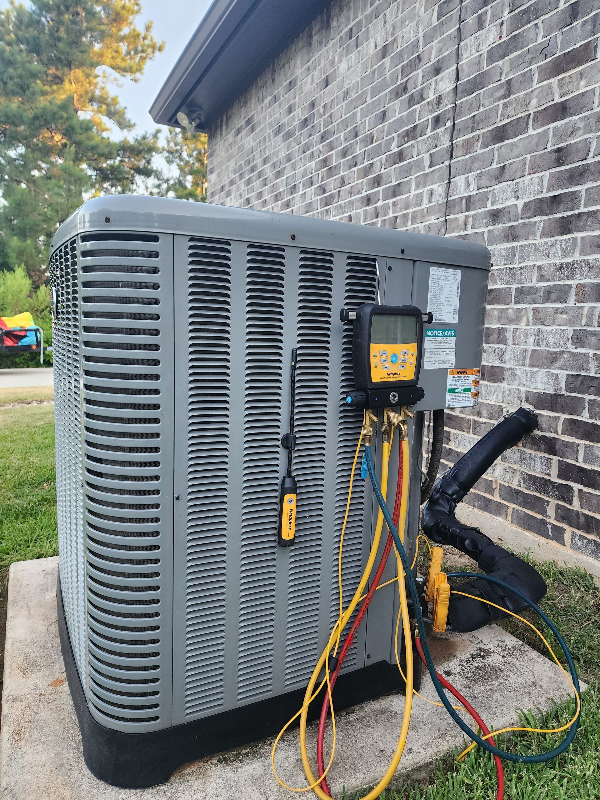 Outdoor HVAC unit with attached gauges and colorful refrigerant hoses beside a brick wall