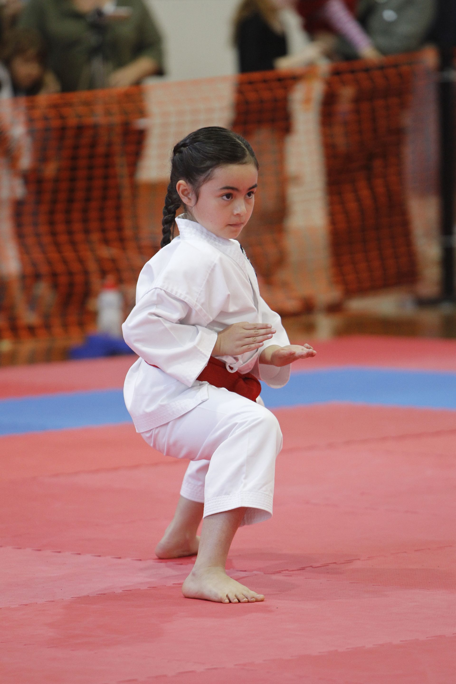 Two martial artists in white uniforms with red belts pose at an indoor competition arena.