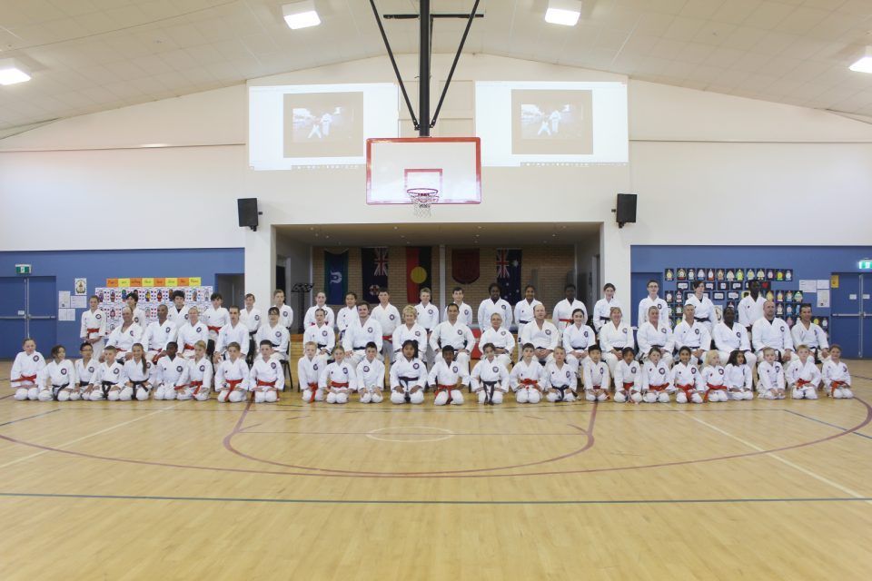 Large martial arts group posed in white uniforms in a gymnasium under a basketball hoop
