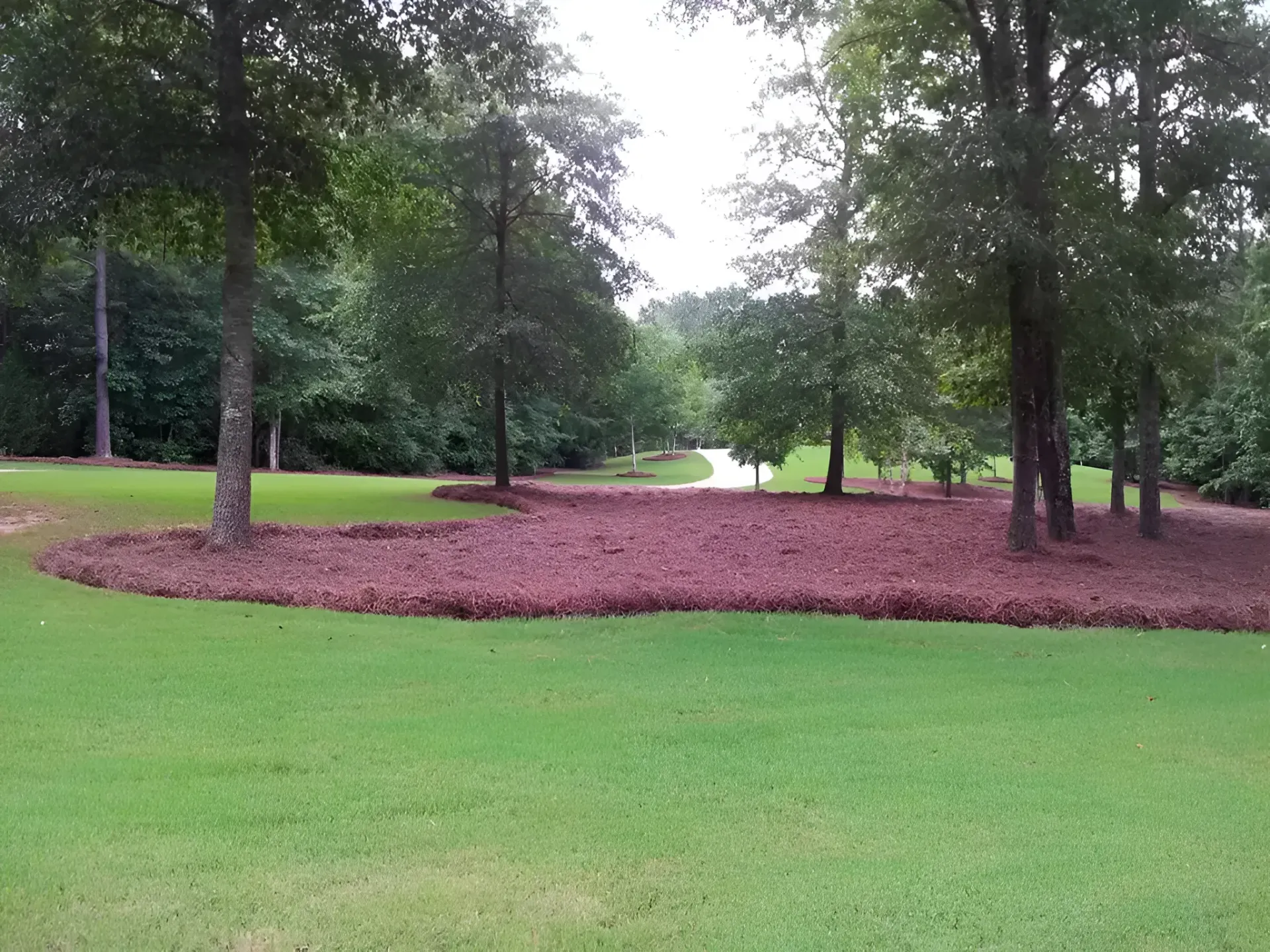 Tree-lined park with green grass and a circular bed of red mulch around a large open area
